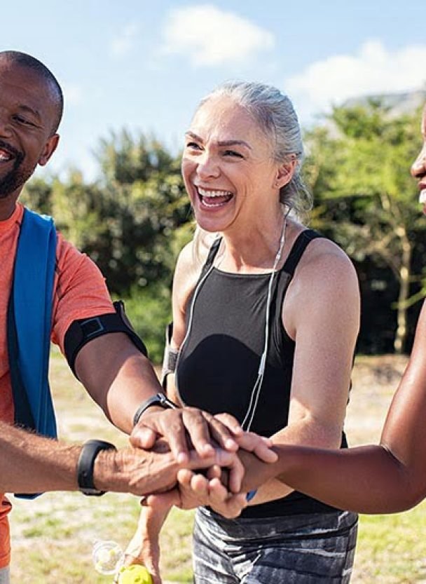 Laughing senior and multiethnic sports people putting hands together at park. Happy group of men and women smiling and stacking hands outdoor. Multiethnic sweaty team cheering after intense training.